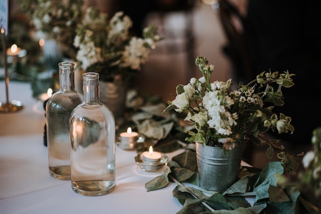 White flowers in a bucket, water bottles and candles on a table decorated with leaves