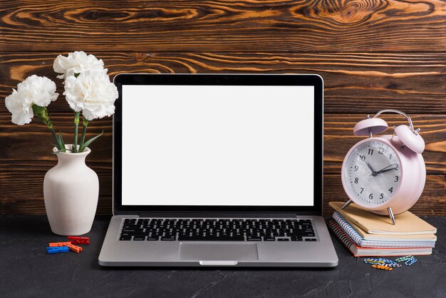 White flowers in the vase; laptop and alarm clock on notebooks against wooden backdrop