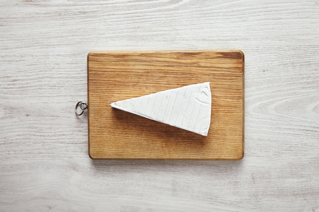 White fresh triangle of tasty brie cheese on cutting board isolated on white aged wooden table in center.