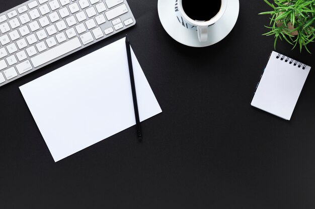 White keyboard; paper; pencil; coffee cup; spiral notepad and pot plant on black background