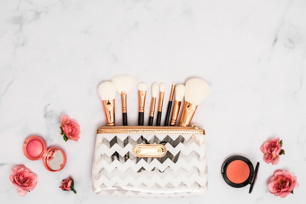 Free photo white makeup bag with brushes; compact powder and roses on textured backdrop