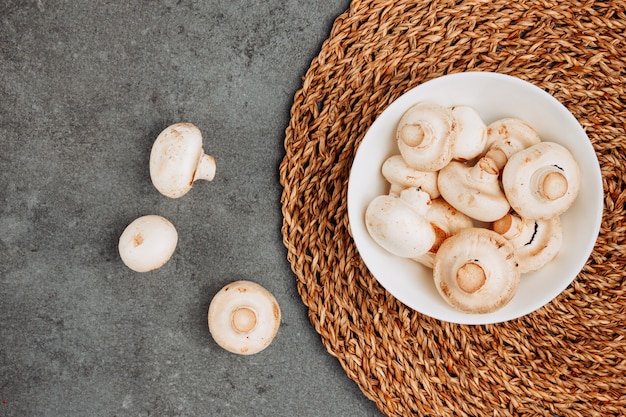 White mushrooms in a bowl on a rattan trivet and gray textured background. top view.