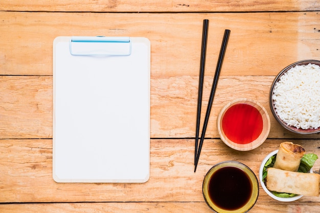 White paper on clipboard near the chopsticks; spring rolls; rice and sauces on wooden desk