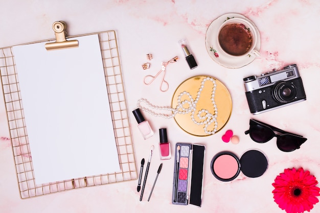 White paper on clipboard; necklace; sunglasses; camera; gerbera flower; coffee cup; necklace and cosmetic products on pink background