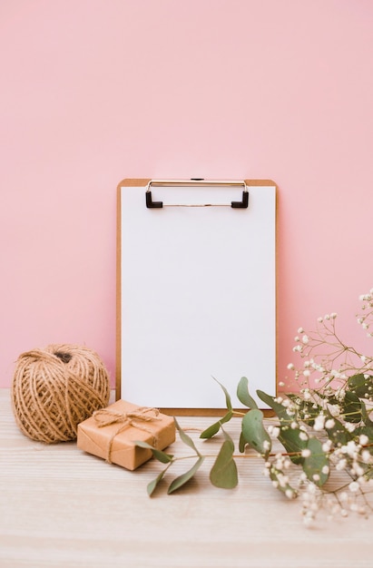 White paper on clipboard with spool; gift box and baby's-breath flowers on wooden desk against pink background