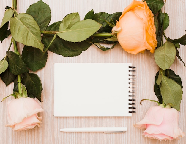 Free photo white pen and blank spiral notepad surrounded with roses over wooden background