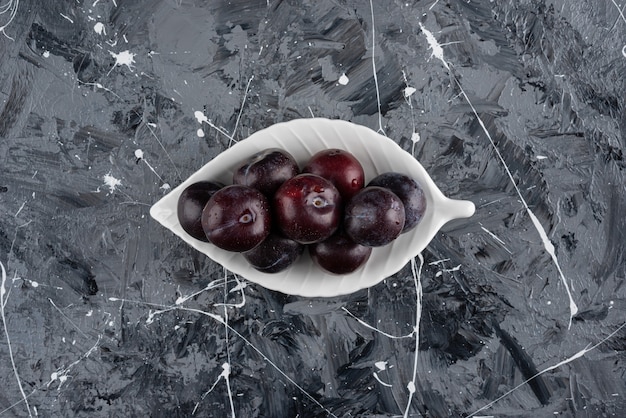 White plate of fresh purple plums on marble surface.