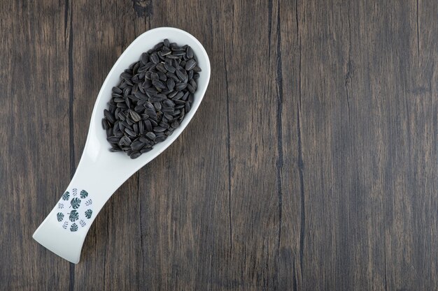 White spoon full of healthy black sunflower seeds placed on a wooden table .