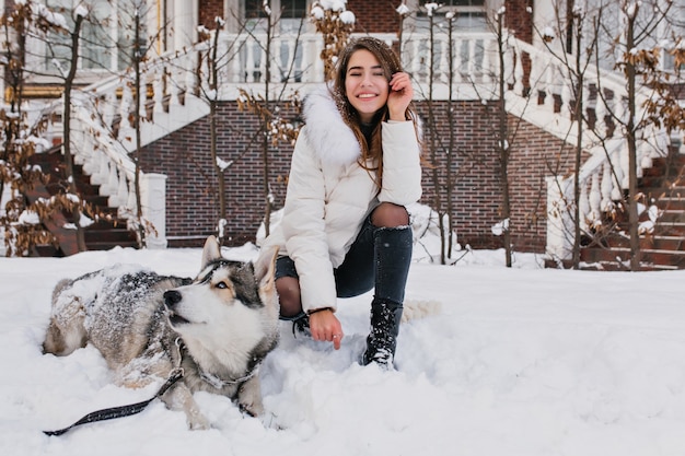 White woman with amazing smile posing with her dog during winter walk in yard. Outdoor photo of cheerful lady wears ripped denim pants sitting on the snow with lazy husky.