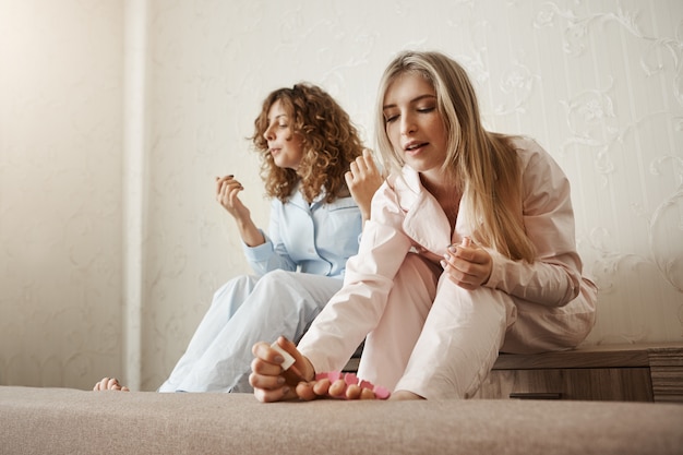 Free photo who needs beauty salons when you can make one at home. portrait of two best friends sitting in room and painting fingernails on feet, wearing nightwear and feeling cozy, having girly party