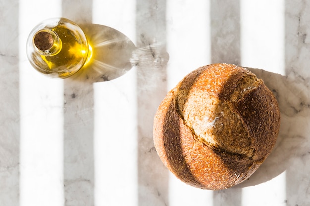 Free photo whole bread and olive bottle on striped shadow over the white marble backdrop