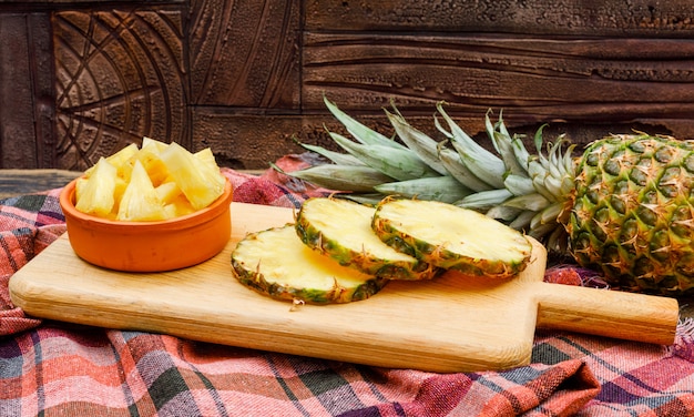 Free photo whole and sliced pineapple in a cutting board and clay bowl on a stone tile and picnic cloth. side view.