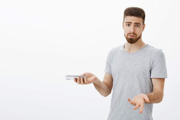 Why keep annoying me I am talking on phone. Confused and bothered silly masculine man with beard and blue eyes shrugging with raised palms holding smartphone, interrupted during important conversation