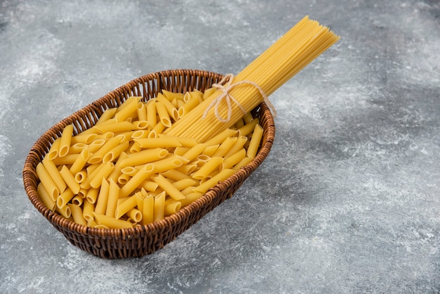 Wicker basket of raw spaghetti and penne pasta on marble surface.