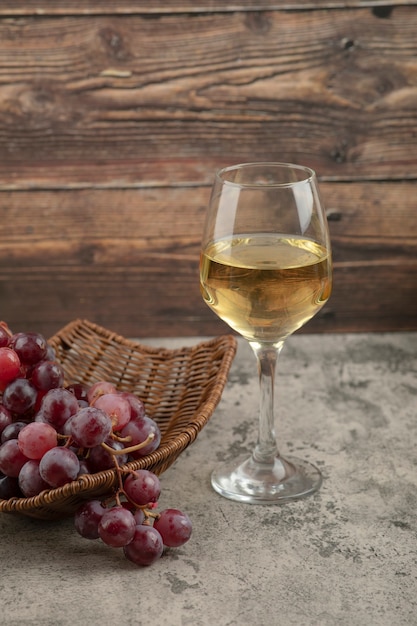 Wicker basket of red grapes with glass of white wine on marble table.