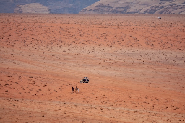 Wide angle shot of two people on camels approaching a car in a desert