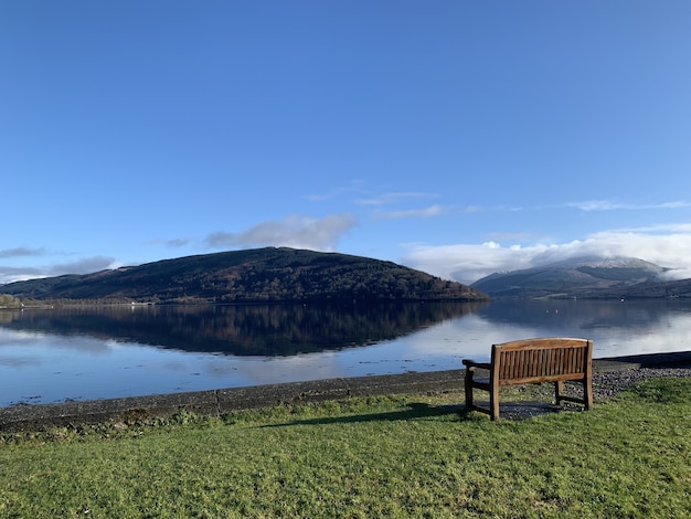 Free photo wide angle shot of a wooden bench on a green field in front of water and a mountain