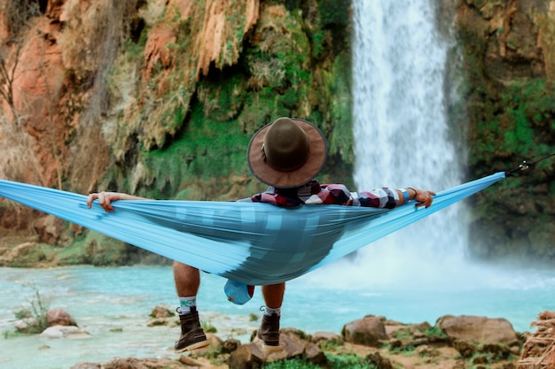 Free Photo wide shot of a male lying on a hammock beside a waterfall flowing down from a hill