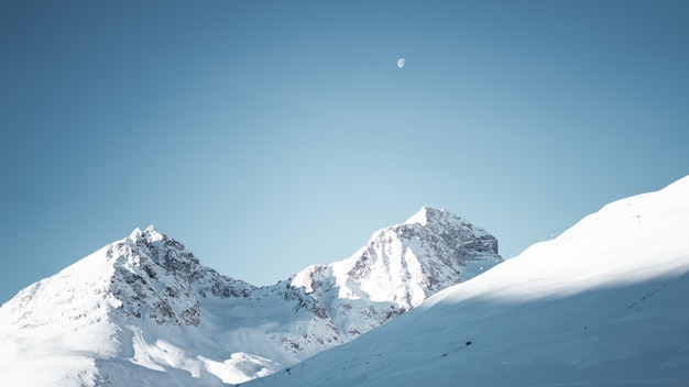 Wide shot of mountains covered in snow under a clear blue sky with a half moon