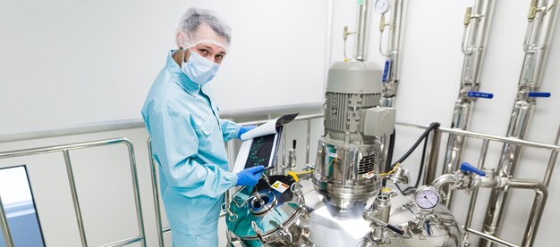 Widescreen picture scientist in blue laboratory suit and gas mask stand in clean room and check graphs in tablet near big steel tank