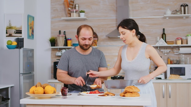 Free Photo wife cooking eggs for husband during breakfast while he is smearing butter on roasted bread. wearing pajamas in the morning, preparing meal together, young happy couple love and marriege