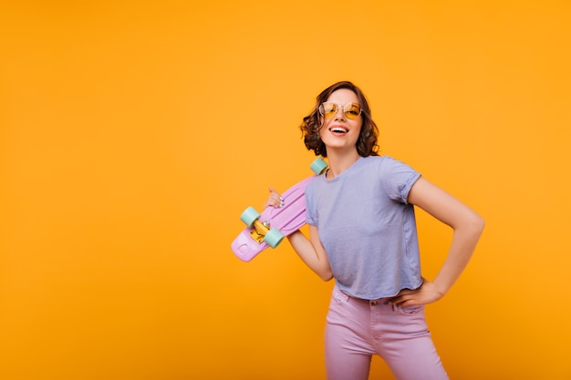 Free photo winsome girl in yellow-colored glasses posing. dark-haired blithesome lady with skateboard standing.