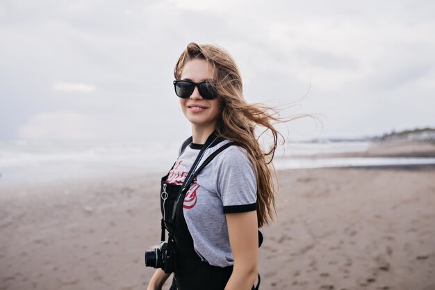 Winsome long-haired girl with camera walking down the sea coast. Outdoor portrait of carefree young woman in dark sunglasses posing.