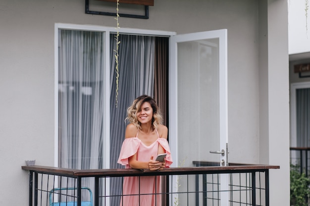 Free Photo winsome tanned girl with phone in hands smiling and looking away. cheerful young lady in pink outfit standing at hotel balcony.