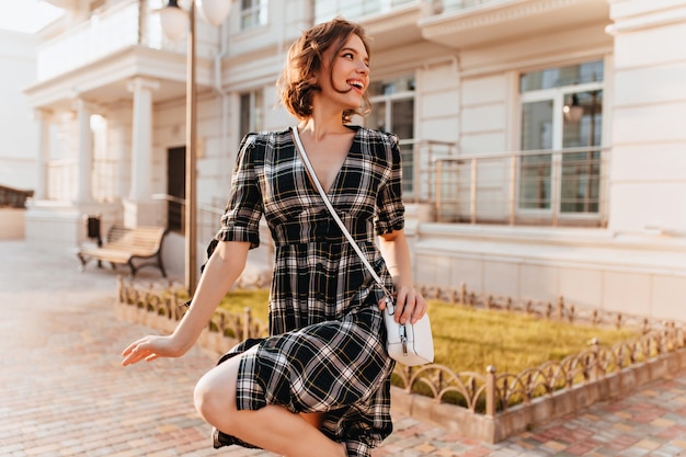 Free Photo winsome white girl in checkered dress walking around in warm autumn day. portrait of spectacular woman with curly brown hair.