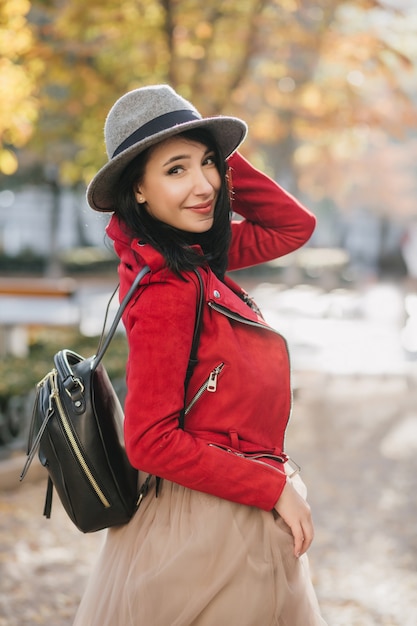 Winsome woman in bright red jacket looking over shoulder during walk in autumn park