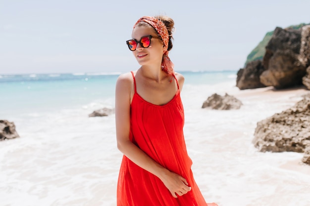 Winsome woman in red dress posing at beach in weekend morning. Carefree caucasian girl in trendy summer accessories standing in the beach