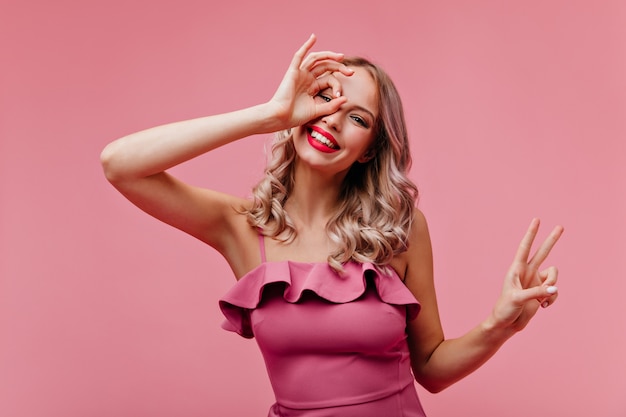 Winsome woman with blonde wavy hair fooling around on rosy wall