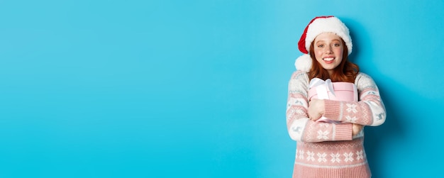 Free Photo winter and celebration concept dreamy redhead girl in santa hat hugging her christmas gift smiling h