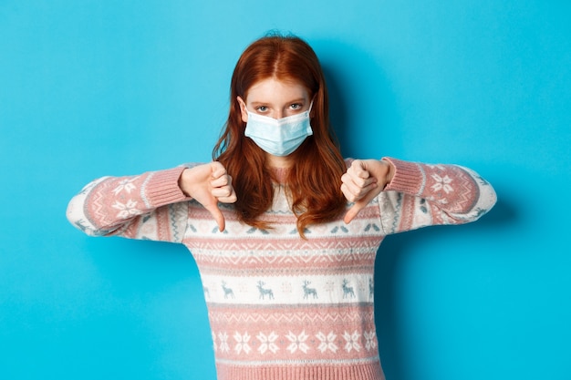 Winter, covid-19 and pandemic concept. Upset and angry redhead girl in face mask showing disapproval, thumbs down in dislike, standing over blue background