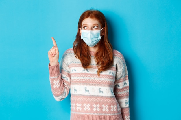 Winter, covid-19 and quarantine concept. Curious redhead girl in medical mask picking product, looking and pointing at upper left corner promo, blue background