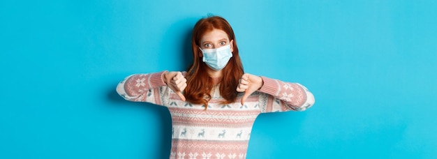 Free photo winter covid and pandemic concept displeased redhead woman in medical mask showing thumbsdown in dis