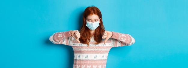 Free Photo winter covid and pandemic concept moody redhead girl in face mask showing thumbsdown disapprove and