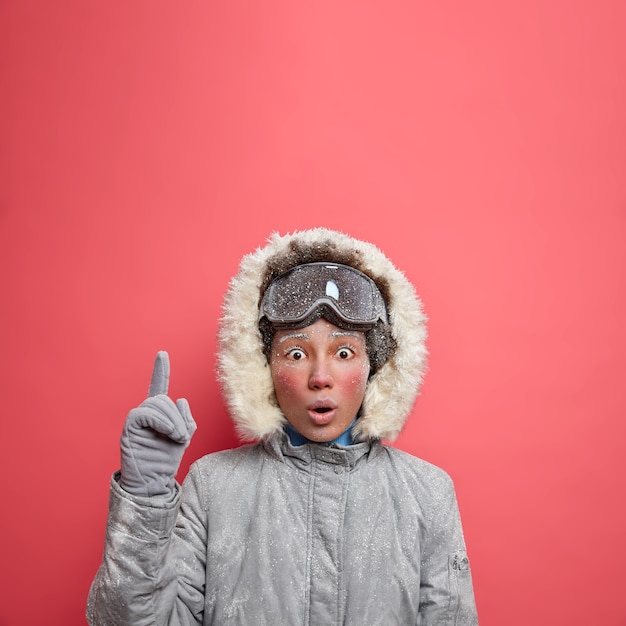 Free photo winter holiday concept. shocked frozen young woman keeps mouth widely opened and points above stunned by very low temperature and heavy snowfall goes skiing during december dressed in outerwear