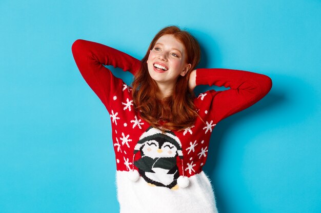 Winter holidays and celebration concept. Relaxed teenage girl holding hands behind head and looking left at promo, resting on Christmas Eve, standing over blue background.