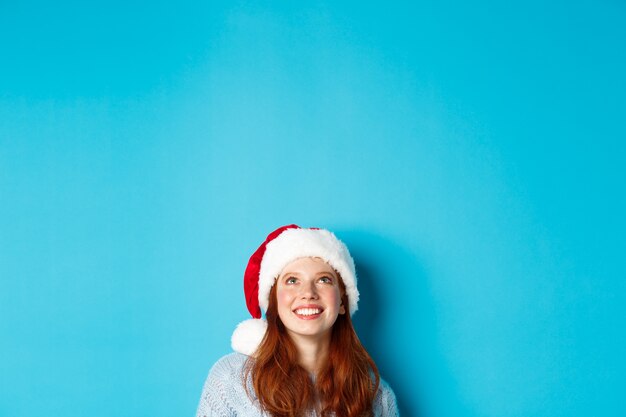 Winter holidays and Christmas eve concept. Head of cute redhead girl in santa hat, appear from bottom and looking up at copy space, staring logo, standing over blue background.