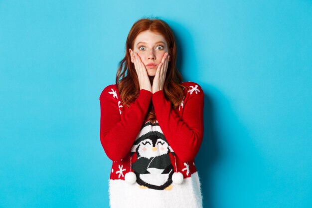 Winter holidays and Christmas Eve concept. Impressed and speechless redhead girl gasping, staring at camera with disbelief, standing in xmas sweater against blue background