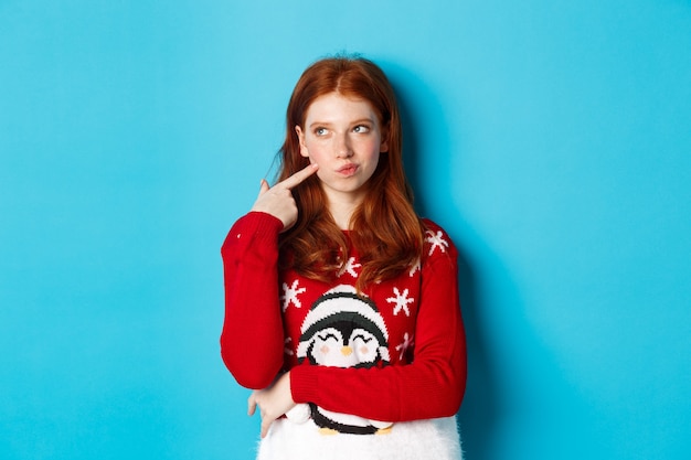 Free Photo winter holidays and christmas eve concept. pretty redhead girl in xmas sweater, touching cheek thoughtful and smiling, making choice, looking at upper left corner and thinking, blue background.