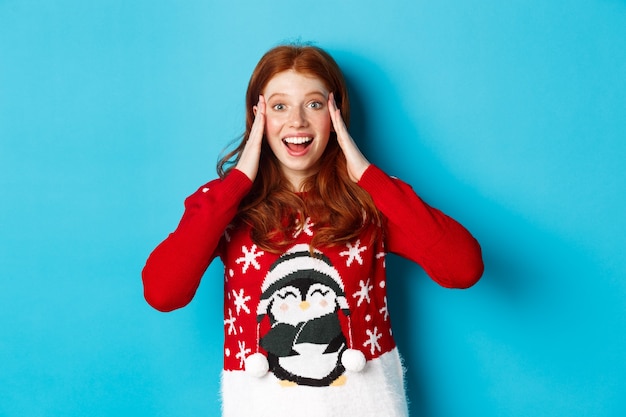 Free photo winter holidays and christmas eve concept. surprised and excited redhead girl, holding hands on head and stare with disbelief at xmas gift, standing over blue background.