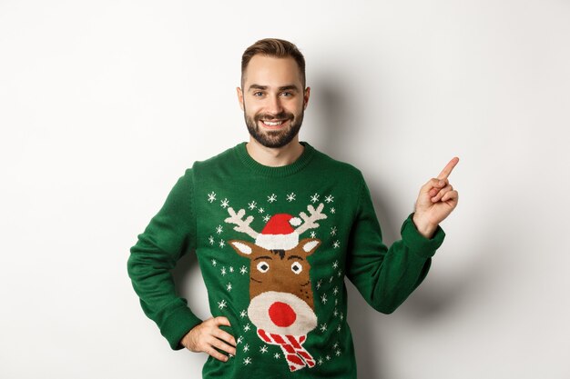 Winter holidays and christmas. Handsome bearded adult man pointing finger right at promo banner, standing in funny sweater against white background