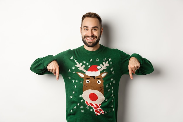 Free photo winter holidays and christmas. handsome smiling man with beard, pointing fingers down and showing promo offer, wearing green sweater, standing over white background.