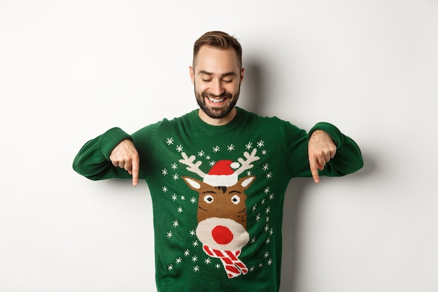 Winter holidays and christmas. Happy caucasian man in green sweater, pointing fingers down and looking satisfied, standing over white background