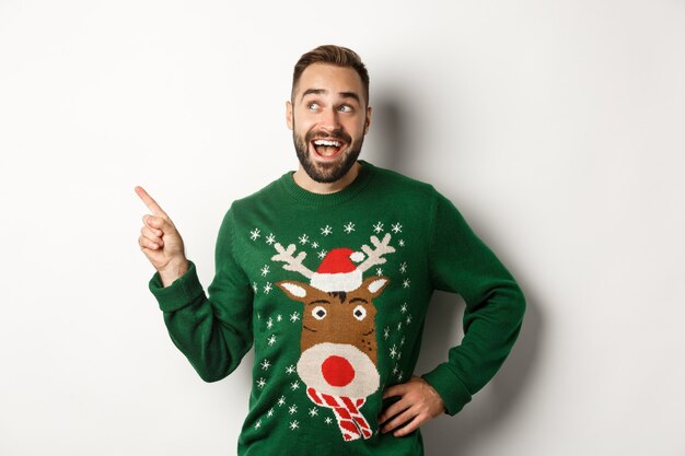 Winter holidays and christmas. Happy man in funny sweater pointing finger at upper left corner logo, showing advertisement, white background.