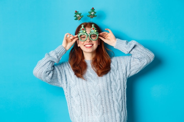 Winter holidays and Christmas sales concept. Beautiful redhead female model celebrating New Year, wearing funny party headband and glasses, smiling silly, blue background