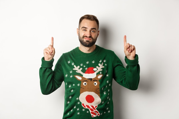 Winter holidays and christmas. Unamused bearded guy in funny sweater pointing fingers up, showing something unpleasant, white background.