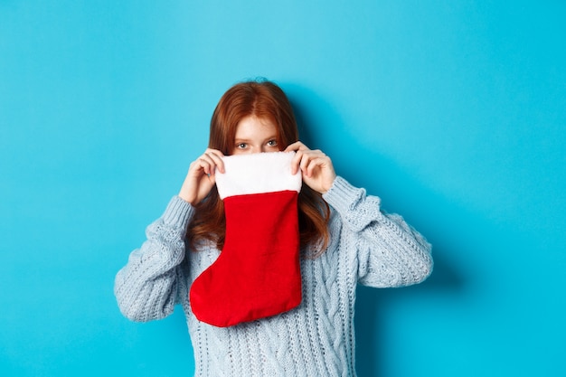 Free photo winter holidays and gifts concept. funny redhead girl looking inside christmas stocking and smiling with eyes, standing against blue background.